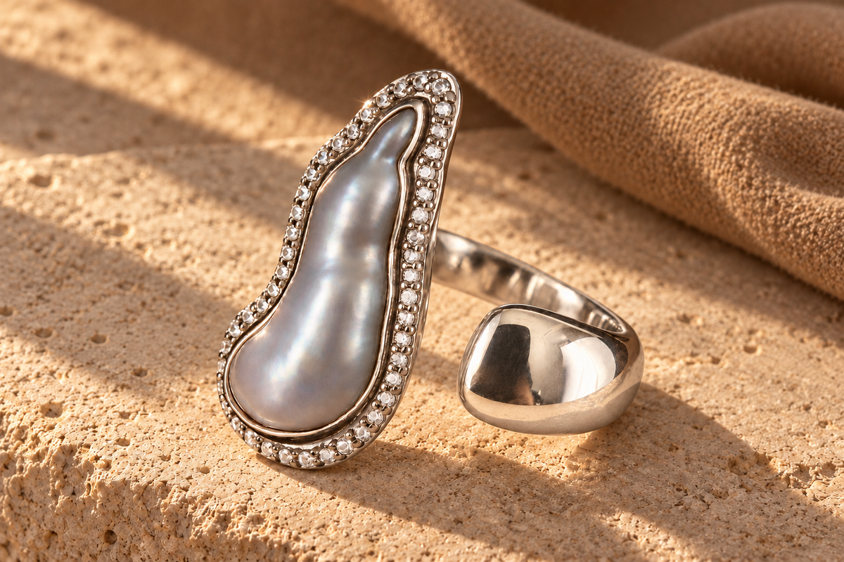 Complete grey baroque pearl open ring on textured stone beside camel fabric in warm golden light.