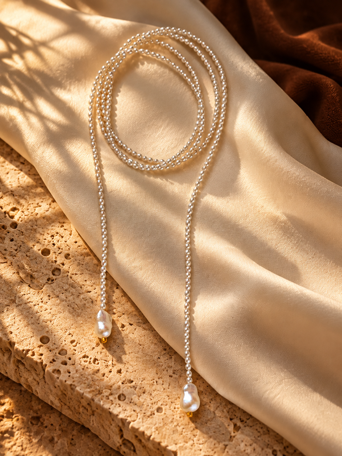 A long pearl lariat with two baroque pearl ends on ivory silk and warm stone in golden sunlight